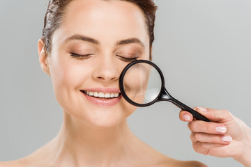 cheerful woman with closed eyes holding magnifying glass near face isolated on grey