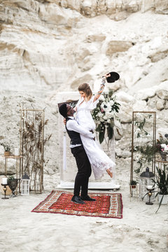 African groom and Caucasian bride embracing each other in a boho style before the wedding arch from fresh flowers. Groom is holding Bride in his arms, smiles and laughs.