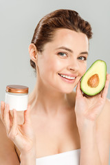 happy woman holding half of avocado and container with cosmetic cream isolated on grey