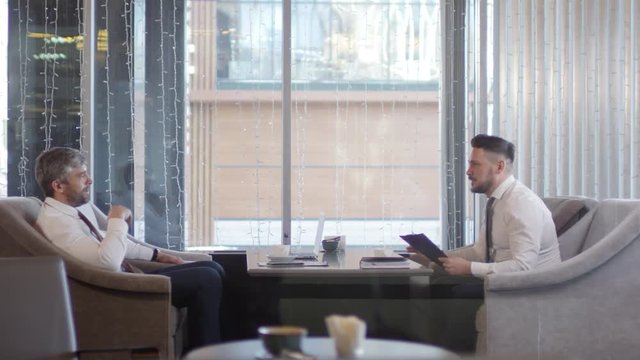 Wide shot of two businessmen sitting in caf&eacute; and communicating. One man answering telephone call and standing up while the other man continuing sitting