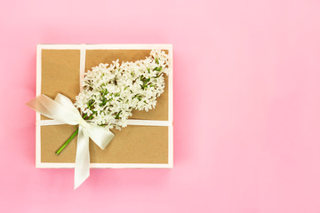 Gift box with holiday bow and white lilac flowers and green leaves on light pink background. Copy space.