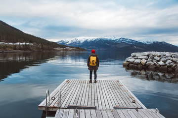 Fototapeta na wymiar Traveler man with a yellow backpack wearing a red hat standing on the background of mountain and lake wooden pier. Travel lifestyle concept. Shoot from the back