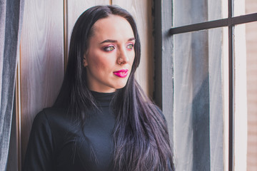 Portrait of a beautiful young brunette girl in a black turtleneck. Sits on the windowsill and looks out the window