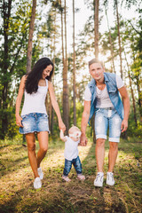 Naklejka na ściany i meble young parents are taught to walk a daughter of one year on nature in a park on a lawn. The first steps of the child's hand with his mother and father