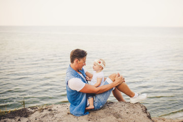 Family summer vacation in nature. father and little daughter sit on sandy cliff for one year with high view of the sea. A man is holding child in his arms. Emotion of happiness, joy and parental love