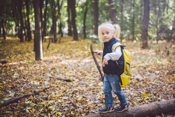 Theme outdoor activities in nature. Funny little Caucasian blonde girl walks walks hiking in the forest on rough terrain with a large backpack. Uses walking stick