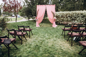 wedding ceremony in nature. arch decorated with a pink cloth. rows of wooden chairs.