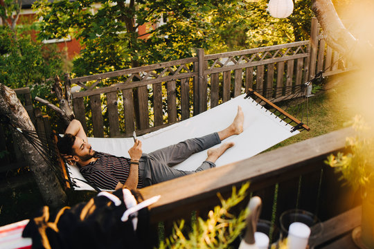 High angle view of young man using mobile phone while lying in hammock at yard