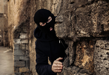 Man in mask behind stone wall watching someone outdoors. Criminal activity