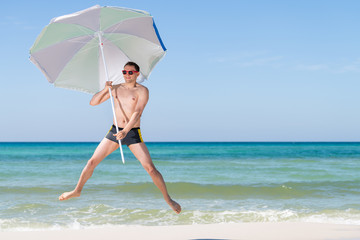 Young attractive fit man happy smiling jumping mid-air on beach on sunny day with red sunglasses in Florida panhandle with ocean holding umbrella