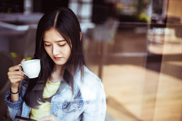 Asian woman with beautiful smile hold mobile phone during rest in coffee shop.Attractive woman drinking coffee.Reflection glass window.