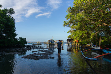 A man was walking in the middle of a mangrove forest whose sea water was receding. In front of him is a Balinese traditional boat view under the blue sky