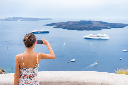 Europe cruise vacation summer travel tourist woman taking picture with phone of Mediterranean Sea in Santorini, Oia, Greece, with cruise ships sailing in ocean background.