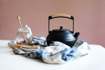Still life about tea time. Black cast iron teapot and jar of honey with spoon are serving on plaid...