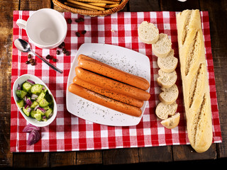 Sausage portion with bread baguette on table setting on checkered tablecloth and cup in farm style. Delightful, unique taste. Homemade, delicious recipes. Top view.