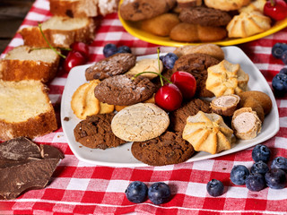 Fototapeta na wymiar Oatmeal Cookies and sand chocolate cake with blueberries on wooden table with gingham cloth in farm style close up. Table setting breakfast for two with cups. High carbohydrate content.