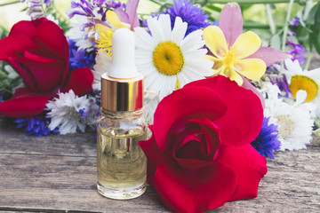 essential oil with a rose in a glass bottle near wildflowers on wooden background.