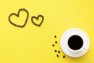 White cup of black coffee on bright yellow table. Two heart shapes created from brown beans. Empty place for nice, lovely, inspirational text, quote or saying.