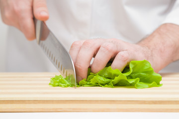 Man's hands cutting green salad leaves with big knife on wooden board. Closeup. Front view. 