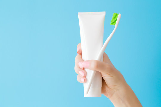Woman's hand holding white tube of toothpaste and toothbrush with green bristles. Pastel blue background. Teeth hygiene. Closeup. Empty place for text or logo.