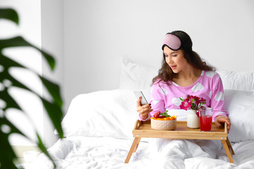 Beautiful young woman with mobile phone having breakfast in bed