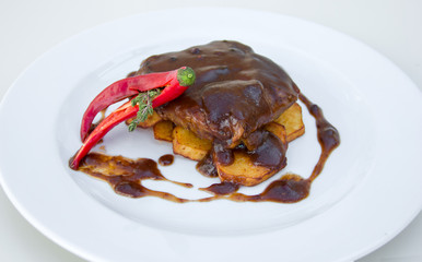 grilled meat and potatoes baked under chocolate sauce, decorated with red pepper, on a white plate, restaurant serve