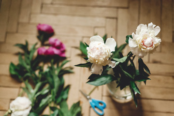 Beautiful pink and white peonies on rustic wooden floor with old glass jar and scissors, top view. Floral decor and arrangement. Gathering flowers. Rural still life, countryside flowers