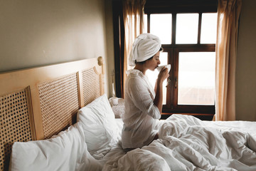 Beautiful happy young woman drinking coffee  or in bed in hotel room or home bedroom. Stylish   brunette girl with white towel enjoying morning in soft light from window