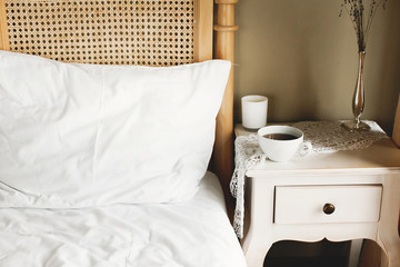 Bed with white sheets and vintage nightstand with white cup of coffee or tea in hotel room or provence home bedroom. Space for text. Enjoying morning concept, coffee in bed.