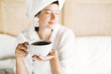 White cup with coffee or tea in girls hands closeup. Beautiful happy young woman drinking tea in bed in hotel room or home bedroom, enjoying morning in soft light. Space for text