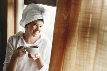 Beautiful happy young woman drinking coffee or tea, sitting at big wooden window in hotel room or home bedroom. Stylish girl in white towel enjoying morning in soft light. Space for text