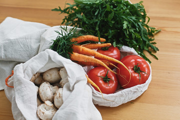 Fresh vegetables in eco cotton bags on wooden table in kitchen. Carrots,tomatoes, arugula, mushrooms from market in canvas reusable bags. Zero waste grocery shopping concept. ban plastic