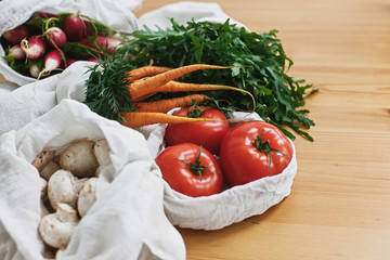 Zero waste grocery shopping concept. Reusable eco cotton bags with fresh vegetables carrots,tomatoes, arugula, mushrooms from market on wooden table. ban plastic. Sustainable lifestyle.