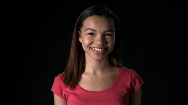 Lockdown of nice African girl wearing pink t-shirt against black background looking at camera and smiling widely