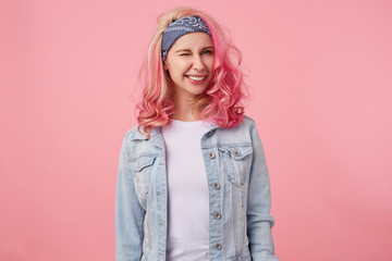 Happy cute smiling lady with pink hair and tattooed hands, looking at the caera and winking, standing over pink background, wearing a white t-shirt and denim jacket.
