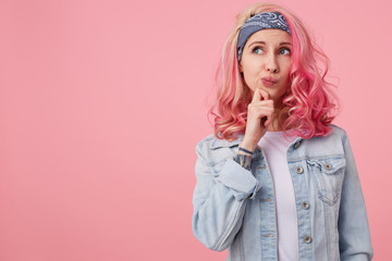 Young thinking cute lady with pink hair, wears a white t-shirt and denim jacket, stands over pink background copy space, looks up thoughtfully and touches the cheek with his finger.