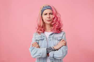 Frowning lady with pink hair and tattooed hand, looking at the camera with disapproval and discontent, standing over pink background with crossed arms, wearing a white t-shirt and denim jacket.