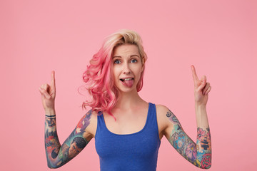 Happy cute lady with pink hair and tattooed hands, standing over pink background, wearing a blue shirt, showing tongue and fooling around. looks up and points fingers at copy space above her head.