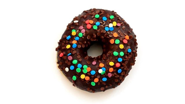 Chocolate donut with colorful sprinkles turns around on white background, top view