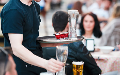 Waiter with tray takes empty beer glasses and brings cocktails to the guests of restaurant on the...