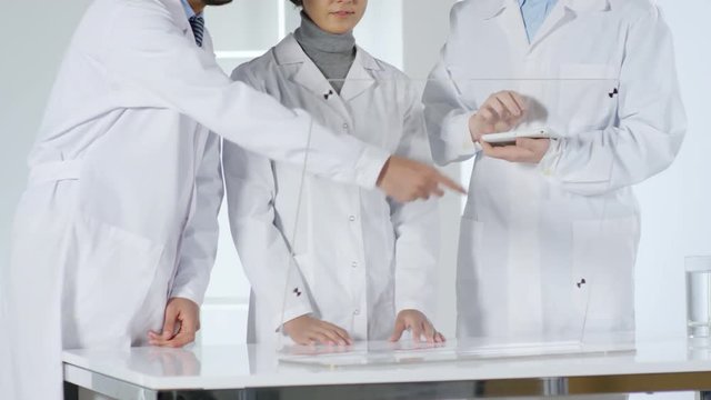 Lockdown of three scientists in lab coats standing together at transparent AR monitor of futuristic computer and having conversation. Video suitable for adding AR graphics
