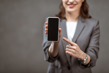 selective focus of businesswoman pointing with finger at smartphone with blank screen on grey