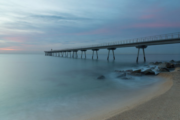 A quiet beach under a cloudy sky at sunrise. Clouds above the pier and at foreground, a long exposure of waves over the rocks and the sand of the beach.