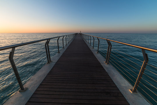 A sunrise seascape with a bridge above the sea with vanishing handrails towards the horizon.