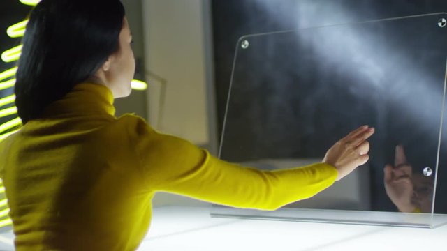 Rare view of young mixed-race woman sitting at glass futuristic computer, tapping and swiping webpages. Video suitable for adding AR graphics