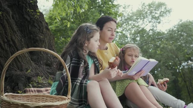Mature grandmother sit in the park near tree on the blanket reading a book with two granddaughters who eatting a tasty bread