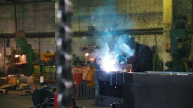 Construction plant. A man in a helmet using a welding machine. A chains hanging on a foreground
