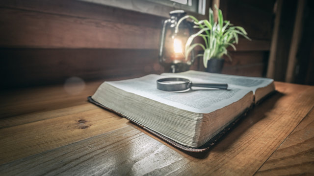 Close Up Bible With Magnifying Glass On Wood Table With Light Of Oil Lamp.