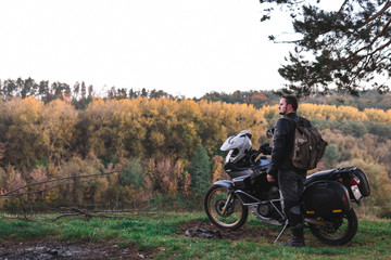 Obraz premium Rider standing with adventure motorcycle, Motorcyclist gear, A motorbike driver looks, concept of active lifestyle, enduro travel road trip. Evening, autumn. Tourist traveler. copy space