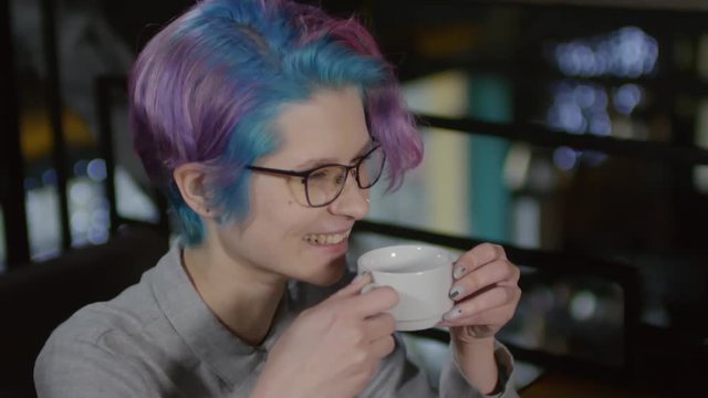 Medium shot of Caucasian hipster-woman drinking tea from tea cup and talking to interlocutor sitting in front of her